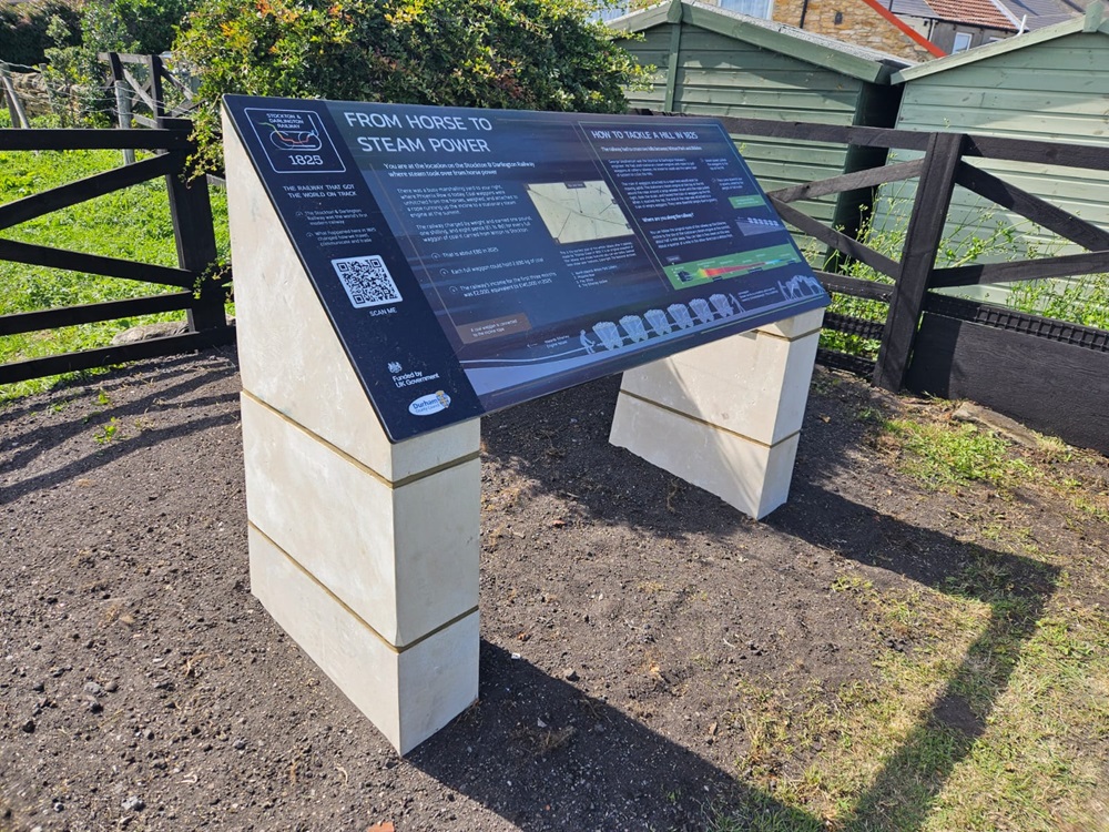View of a interpretation panel that has been installed on the new route. The panel is blue and it is raised on white brick legs set into the ground off the path.