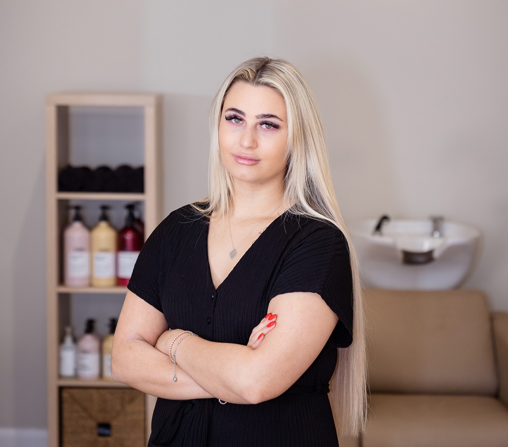 Natalie Lowe owner of Halo Salon standing cross armed looking at camera, with hairdresser sink and accessories in background