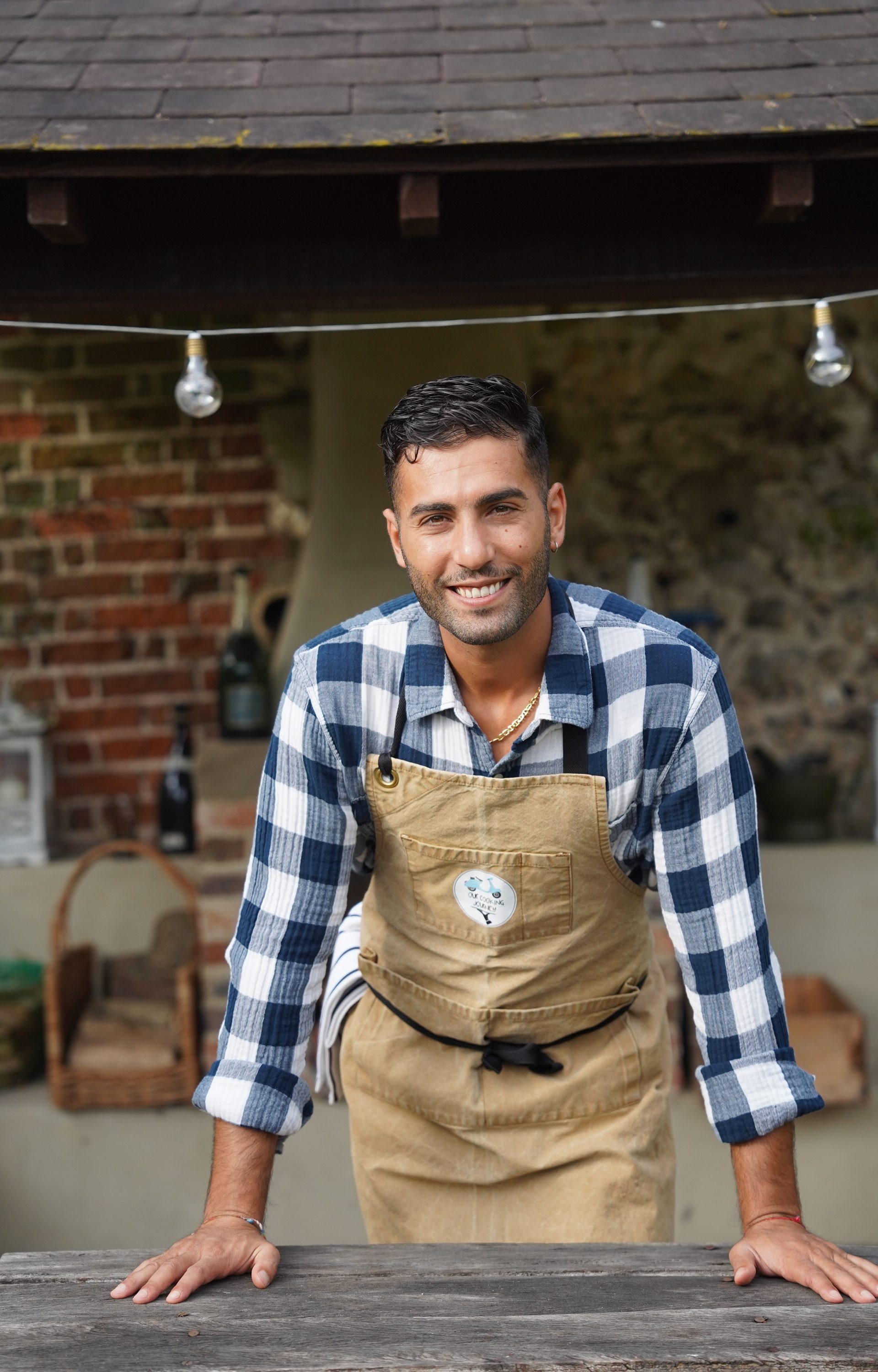 Francesco Mattana chef leaning forward looking into camera with hands resting on wooden bench. Wearing brown apron and blue and white checked shirt
