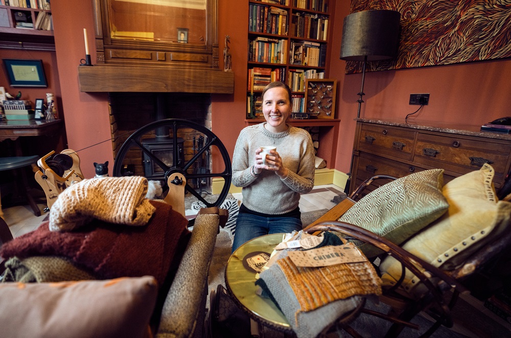 Lauren Leigh Hinthorne sitting in her sitting room surrounded by cushions and sofas holding a mug of tea. to her left is a wool spinning wheel