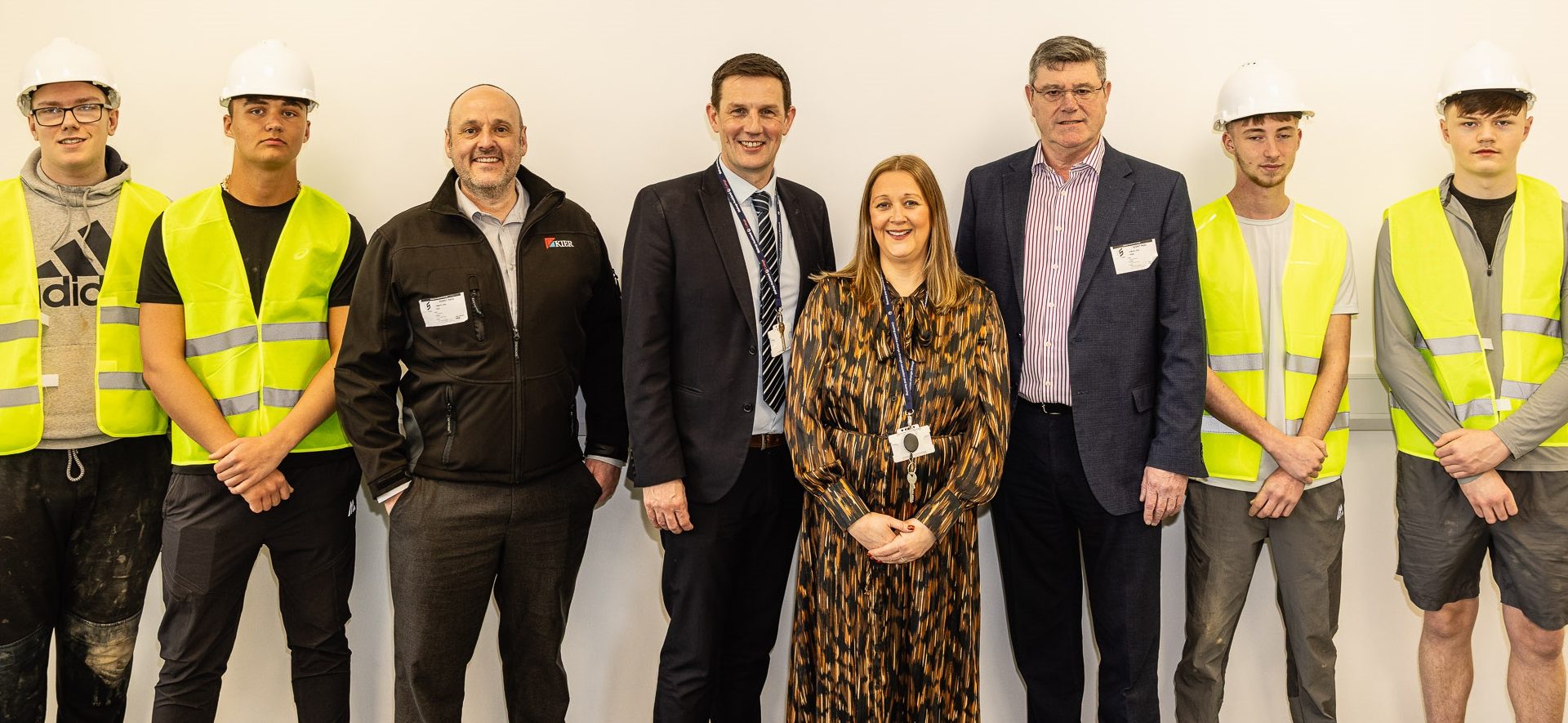 Two bishop auckland students wearing high vis vests and hard hats are standing with college employees and Kier management