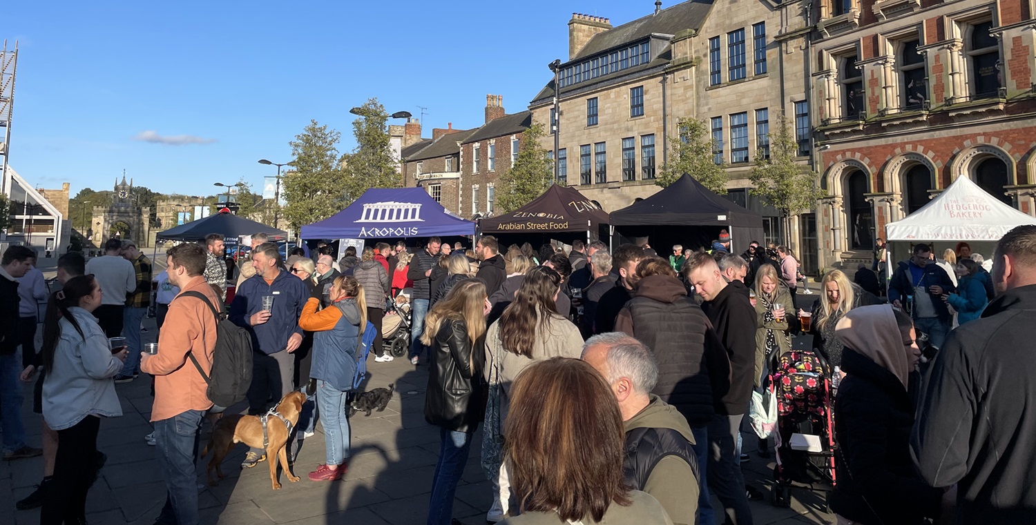Crowd of people in the foreground within the Market Place at Bishop Auckland with a number of market stalls in the background