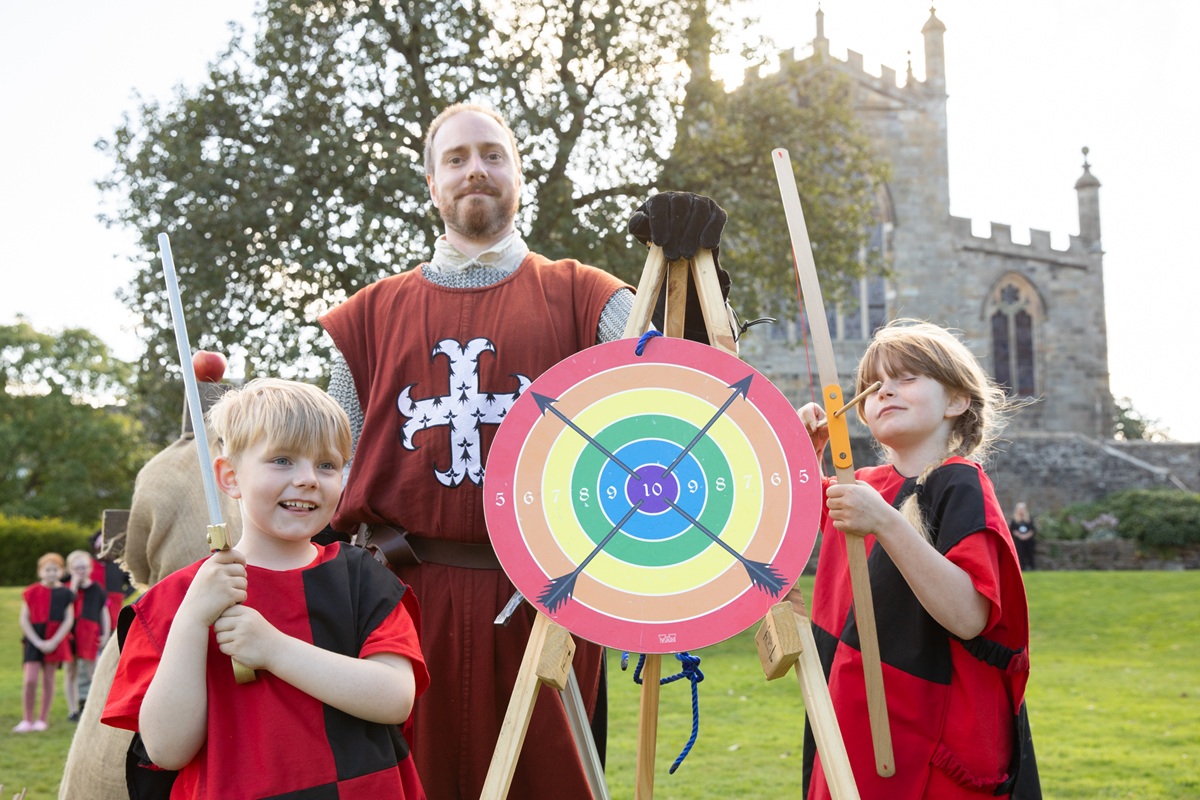 Two children dressed in medieval clothing stand side on either side of a archery board. The adult in the background is wearing medieval clothing