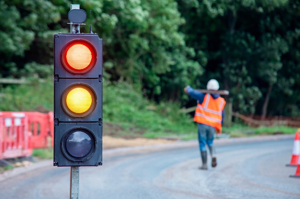 Temporary traffic lights showing red and amber lights. Workman in background with orange high vis jacket carrying wood on his shoulder.