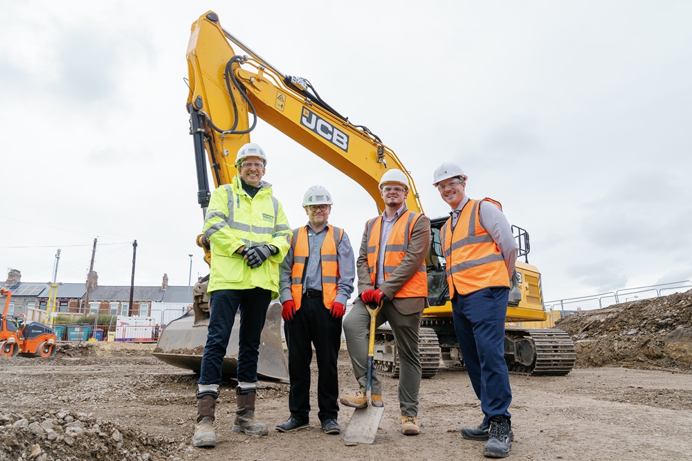 Image of 4 construction workers all wearing high vis jackets and hard hats. One holding a spade, on site with a digger in back ground